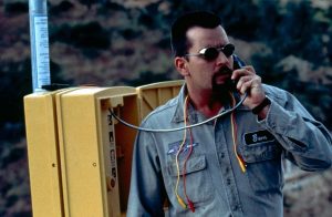 A man in work clothes and sunglasses uses a roadside emergency phone, holding exposed wires; a rocky landscape is in the background.