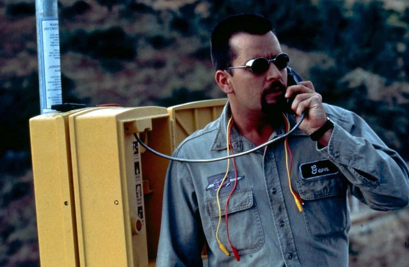 A man in work clothes and sunglasses uses a roadside emergency phone, holding exposed wires; a rocky landscape is in the background.
