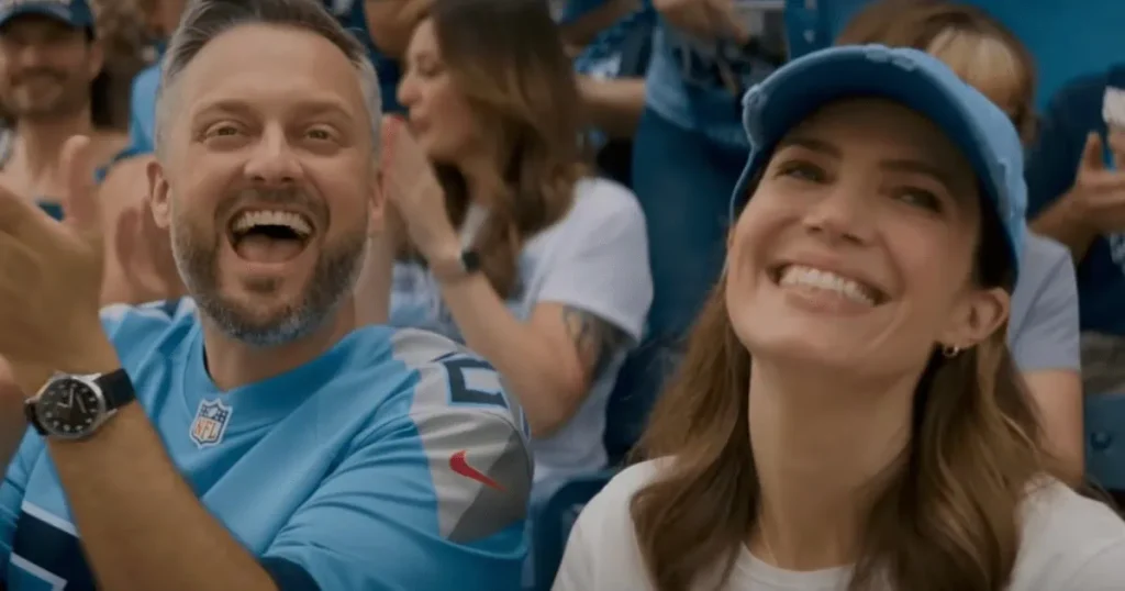 Two people wearing blue sports jerseys and caps are smiling and cheering in a crowded stadium.