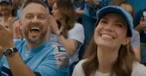Two people wearing blue sports jerseys and caps are smiling and cheering in a crowded stadium.