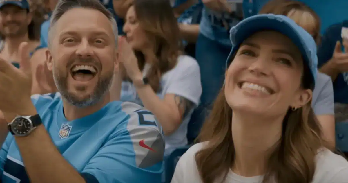 Two people wearing blue sports jerseys and caps are smiling and cheering in a crowded stadium.