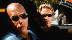Two men wearing sunglasses sit in a car with the top down, looking forward. Trees and a fence are visible in the background.
