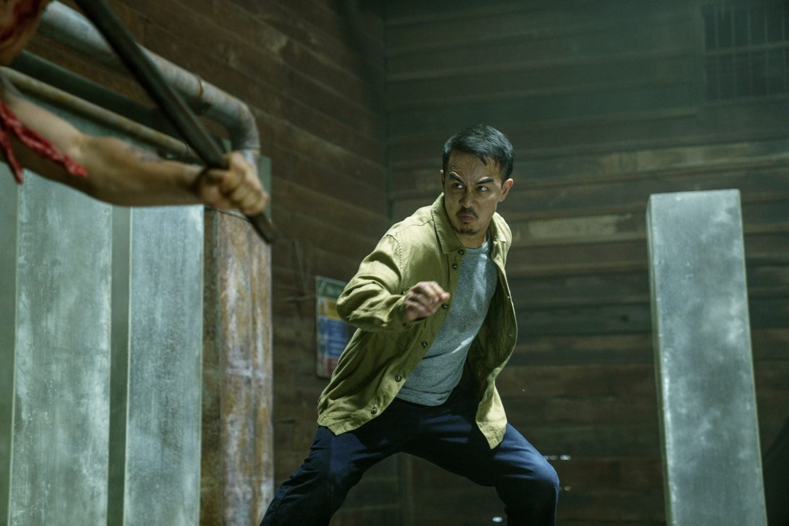 A man in a tan jacket prepares to fight, facing an opponent holding a metal weapon in an industrial, dimly lit room.