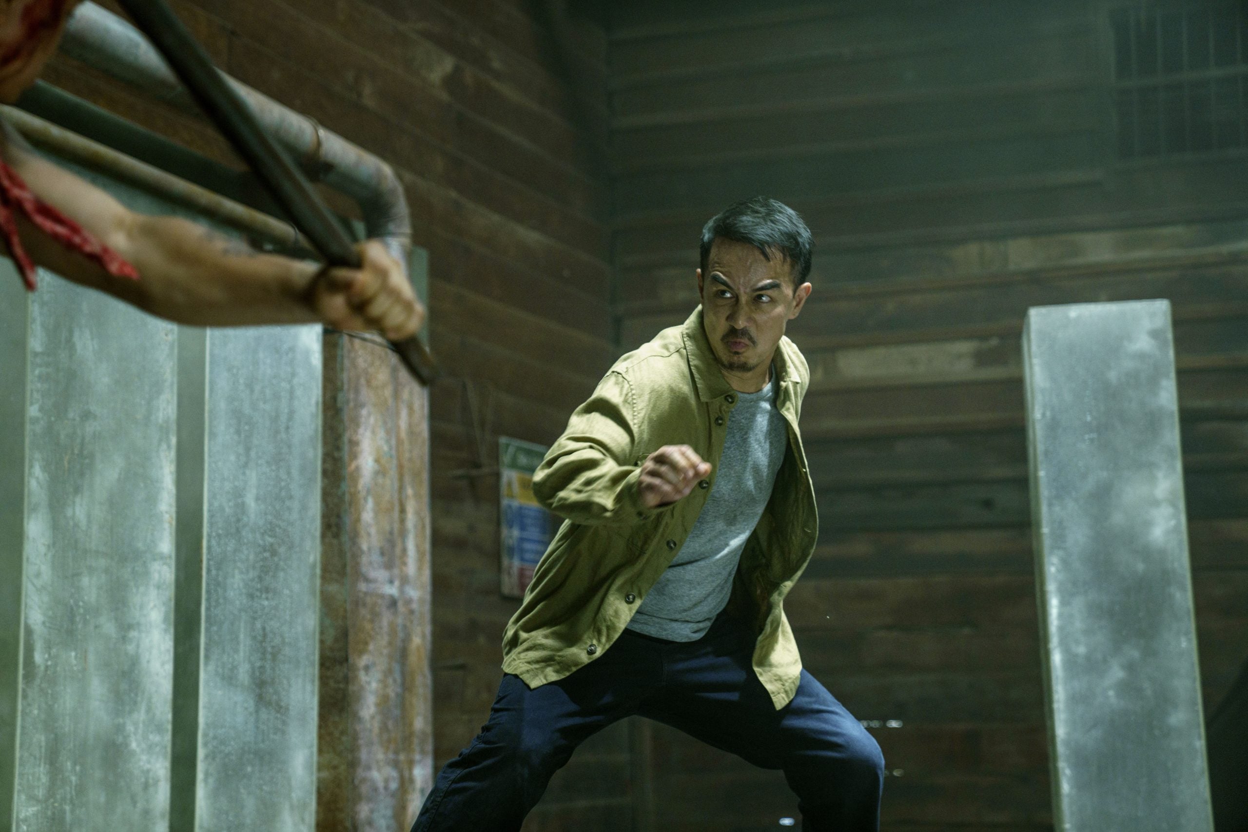 A man in a tan jacket prepares to fight, facing an opponent holding a metal weapon in an industrial, dimly lit room.