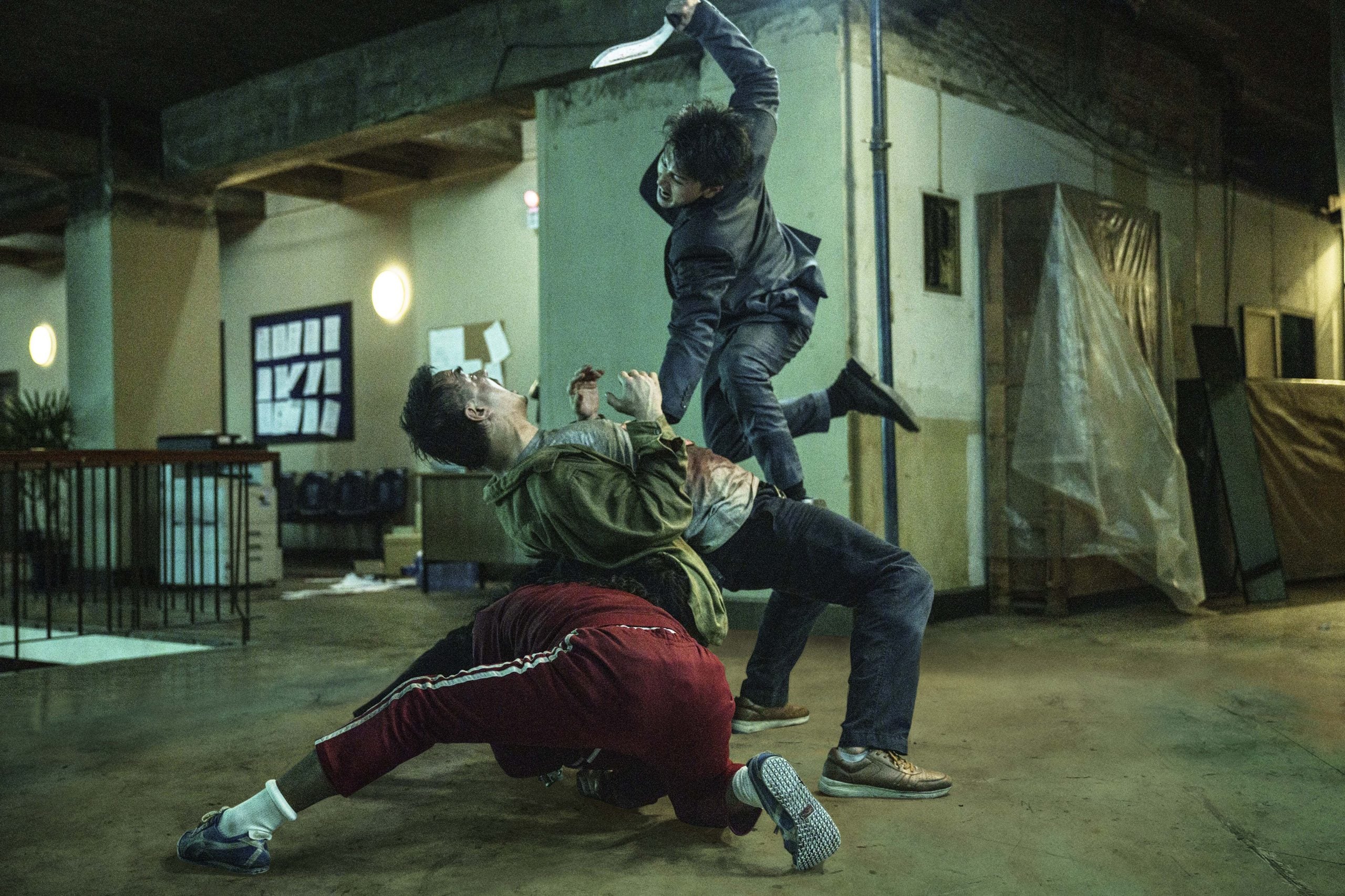 Three people fight in an industrial room; one person in a suit leaps with an object raised, while two others, one in red pants, struggle on the floor.
