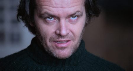 A man with tousled dark hair and stubble, wearing a dark turtleneck sweater, looks upward with an intense, slightly unsettling expression.