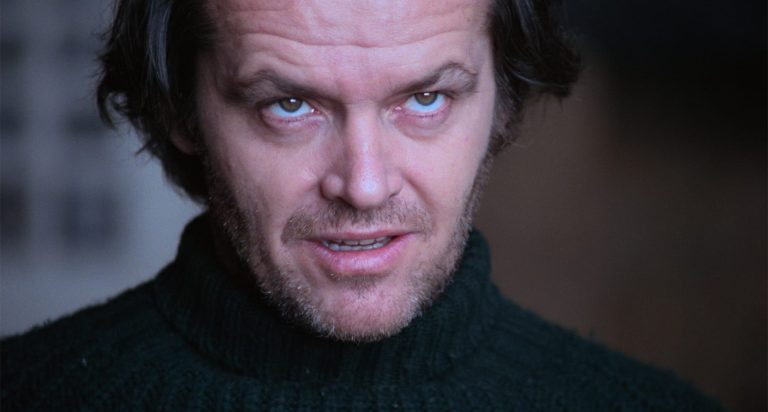 A man with tousled dark hair and stubble, wearing a dark turtleneck sweater, looks upward with an intense, slightly unsettling expression.
