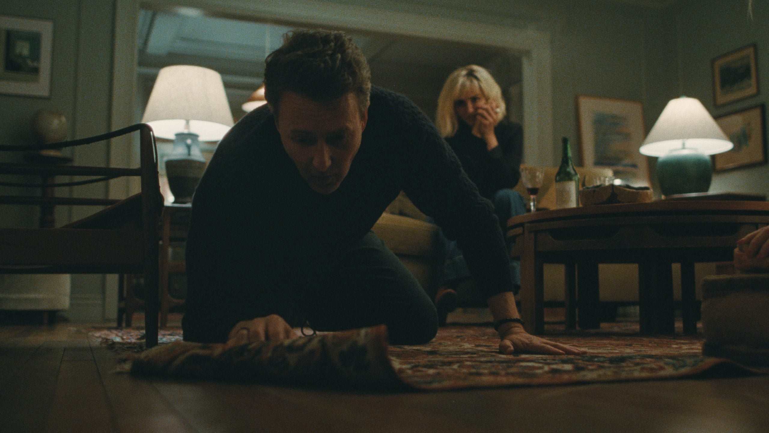 A man kneels on the floor, lifting a corner of a rug, while a woman sits behind him at a table, her hand covering her mouth. The room is dimly lit with lamps.