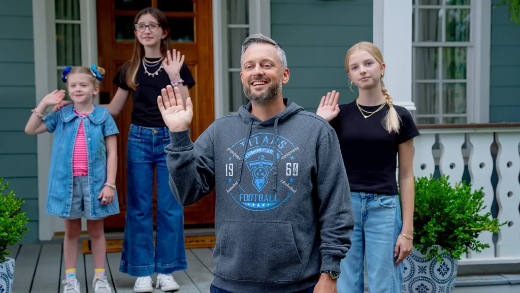 A man and three girls stand on a porch, all waving at the camera. The man is in front, and the girls are behind him, each dressed casually in jeans and t-shirts.
