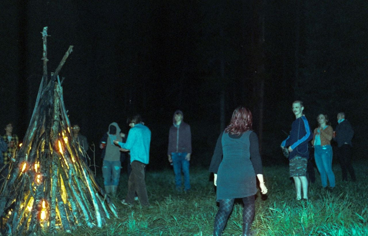 A group of people stand around a bonfire made of tall wooden logs at night in a grassy outdoor area.