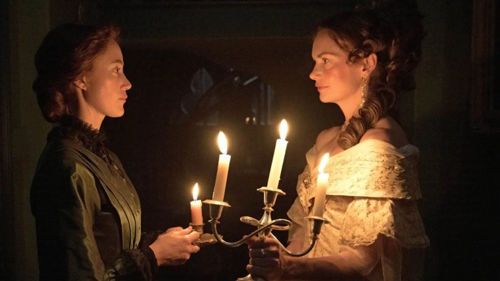 Two women in period clothing stand facing each other in dim light, one holding a candelabra with three lit candles.