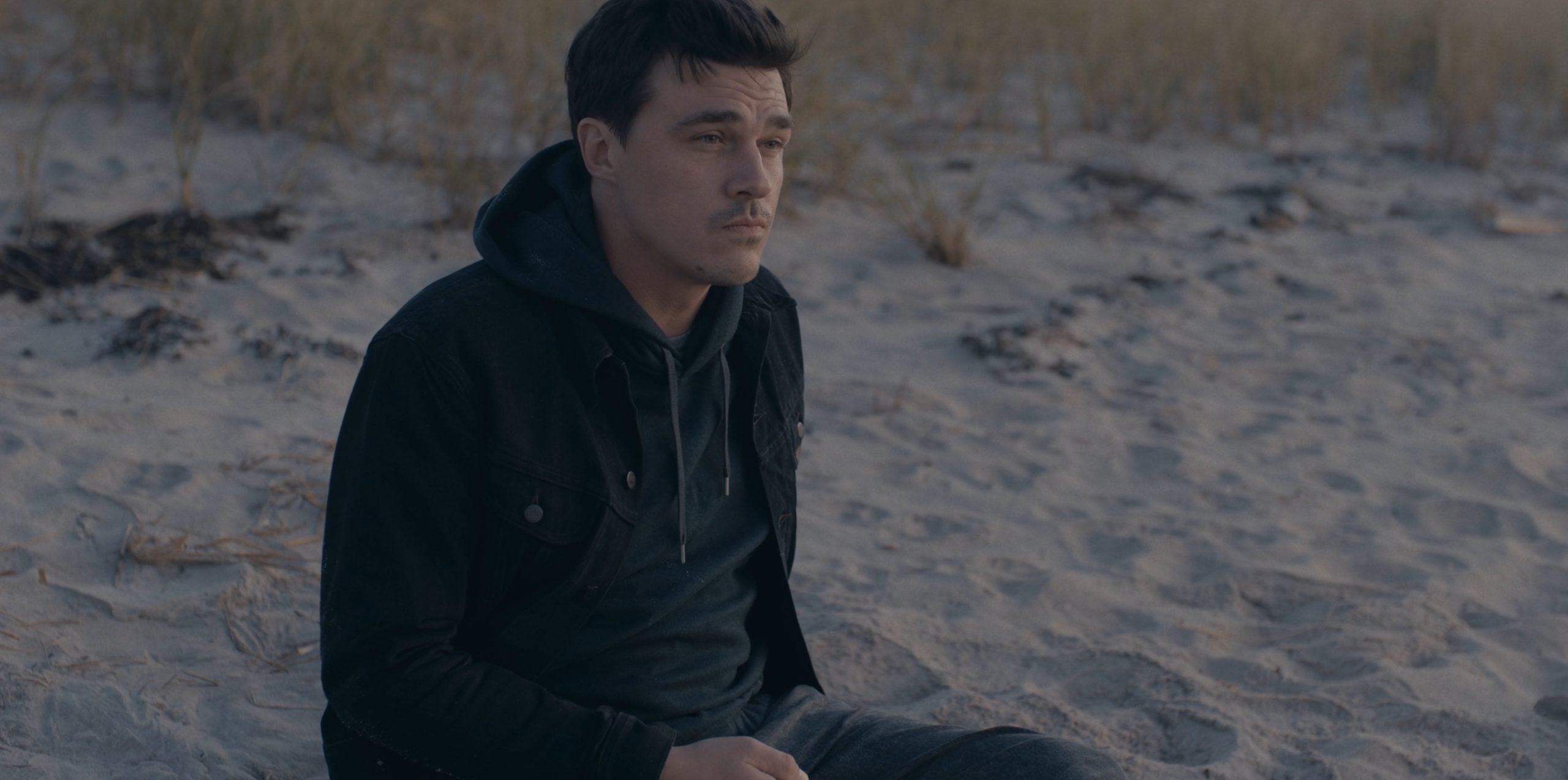 A man in a black jacket and hoodie sits alone on a sandy beach with dry grass in the background, looking off into the distance.