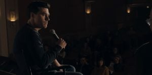 ‘Westhampton’ Review – A Clever, Yet Sluggish Story Of Grief And Acceptance A man sits on stage holding a microphone, facing an audience seated in a dimly lit theater.