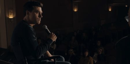 A man sits on stage holding a microphone, facing an audience seated in a dimly lit theater.