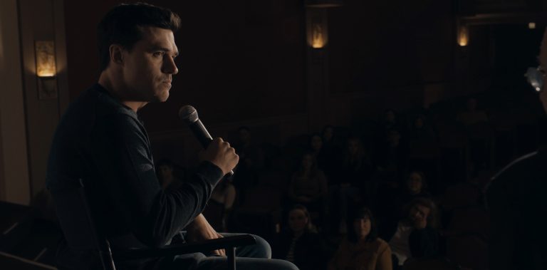 A man sits on stage holding a microphone, facing an audience seated in a dimly lit theater.