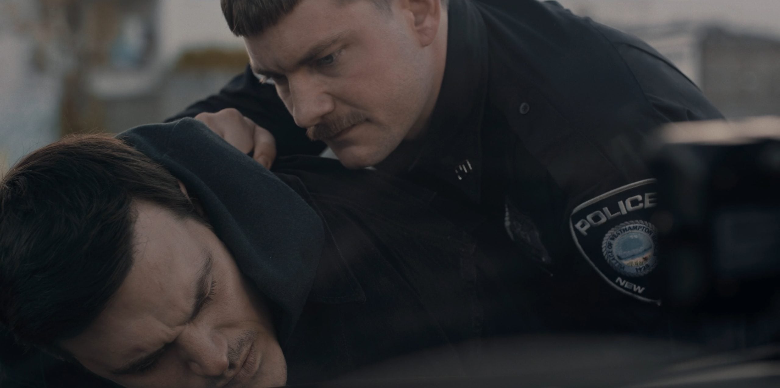 A police officer restrains a man by pressing him against a surface, holding his shoulder while the man winces in pain.
