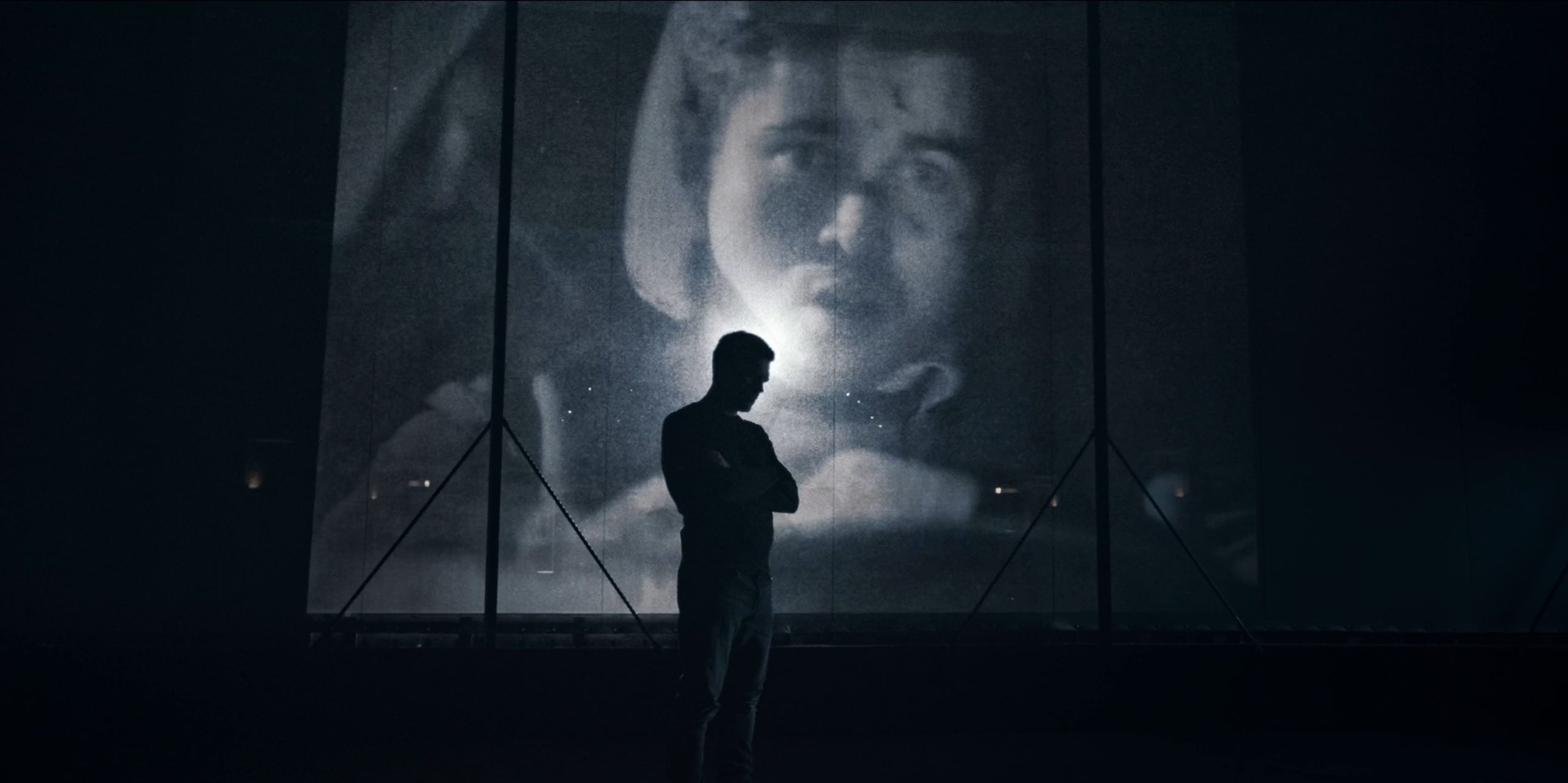 A person stands in silhouette before a large, illuminated screen displaying a close-up black-and-white image of a face.