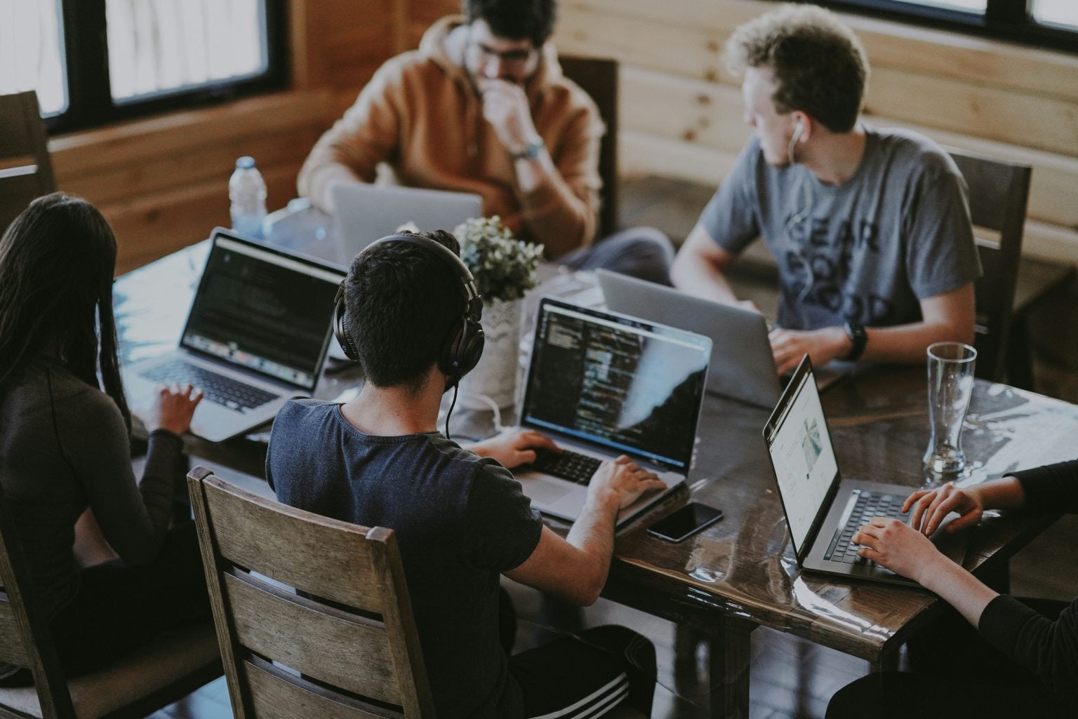 Five people sit around a table working on laptops; one wears headphones and another looks at their phone. The setting appears to be a casual workspace.