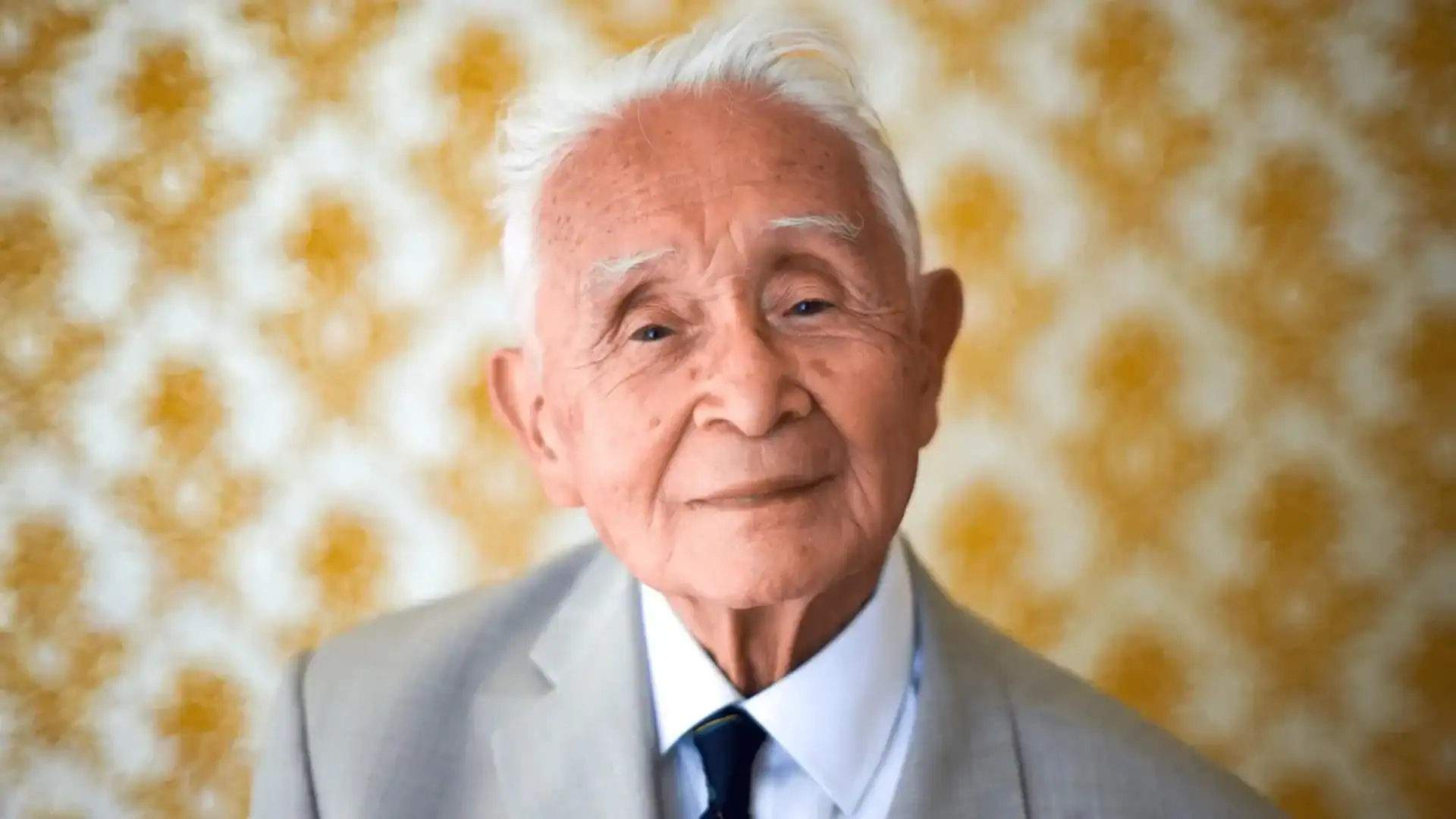 Elderly man with white hair wearing a light gray suit, white shirt, and dark tie, standing in front of a yellow patterned background.