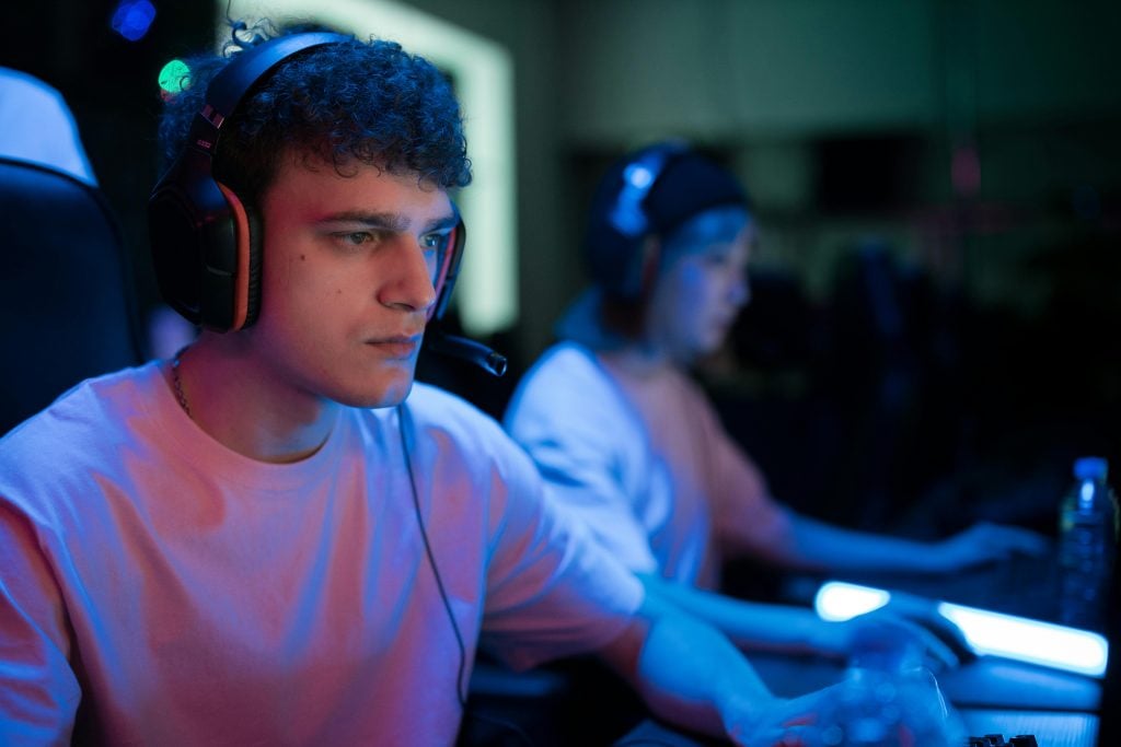 Two young people wearing headsets sit at computers, focused on playing a video game in a dimly lit room with blue lighting.
