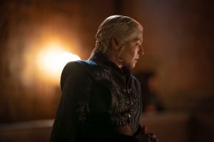 A person with platinum blond braided hair and dark medieval-style clothing stands in profile, facing right, with warm light illuminating the background.