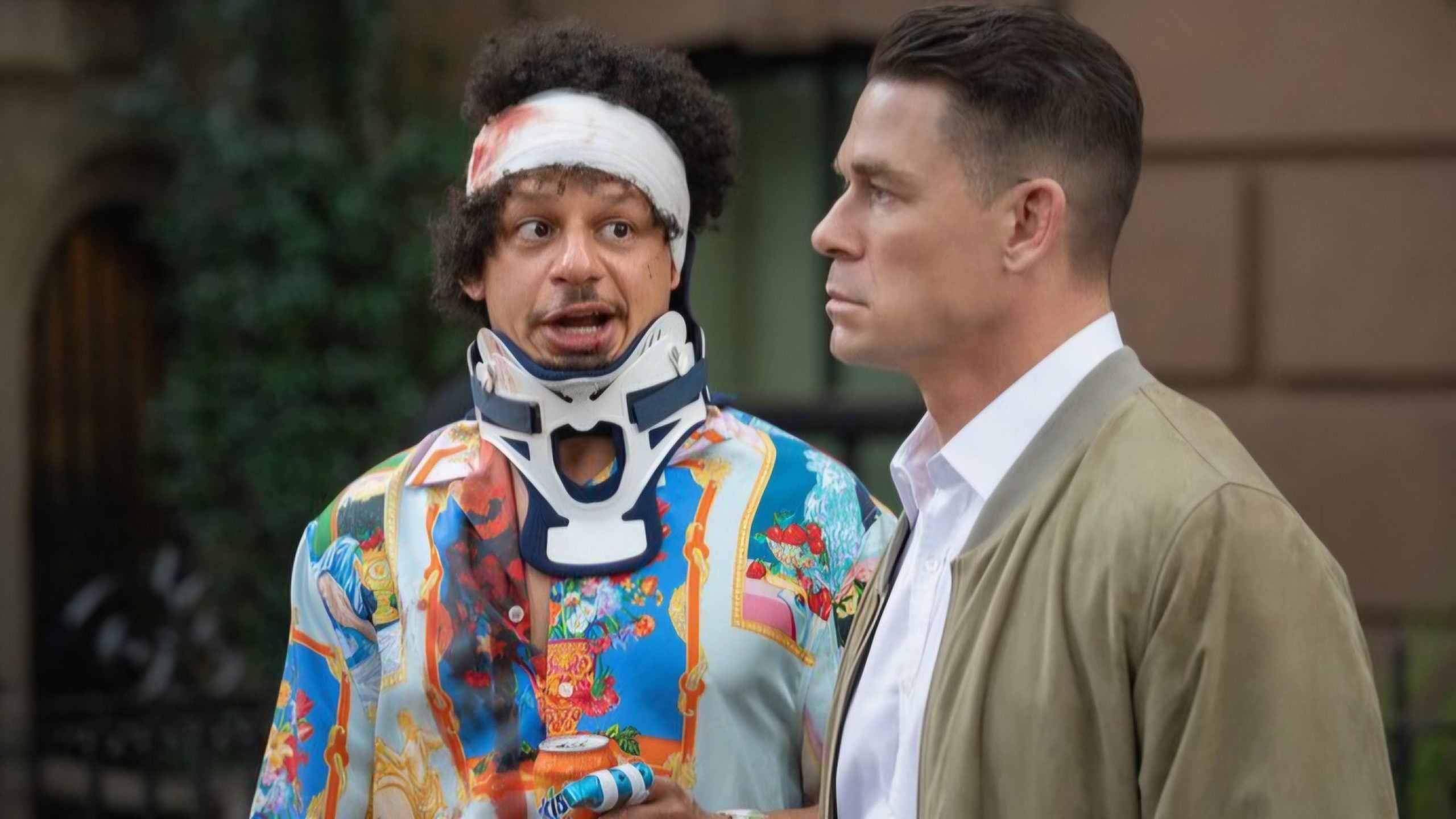 Two men stand outside; one wears a colorful shirt, neck brace, and head bandage, while the other wears a tan jacket and looks serious.
