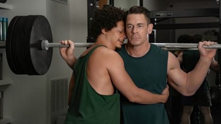 Two men in sleeveless shirts stand in a gym; one is holding a loaded barbell on his shoulders while the other hugs him from the side with a distressed expression.