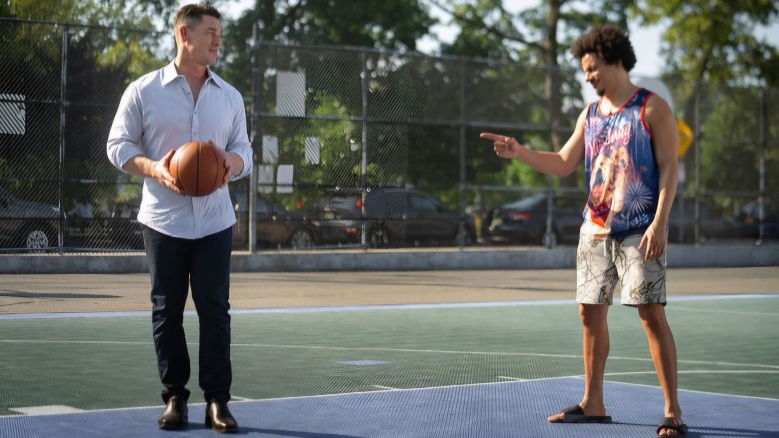 Two men stand on an outdoor basketball court; one holds a basketball and wears a dress shirt and pants, while the other points at him, dressed casually in a tank top, shorts, and sandals.