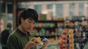 A person stands in a store aisle, holding and comparing two packaged food items while looking at them closely. Shelves with various products are visible in the background.