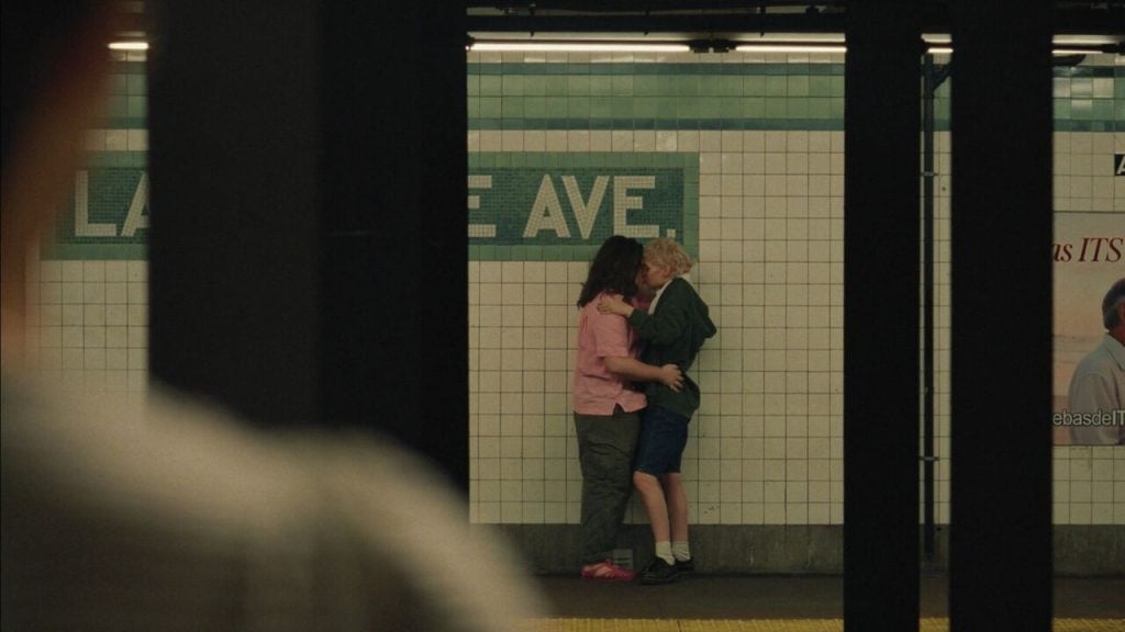 Two people stand closely together, embracing and kissing on a subway platform in front of a tiled wall that reads "AVE.