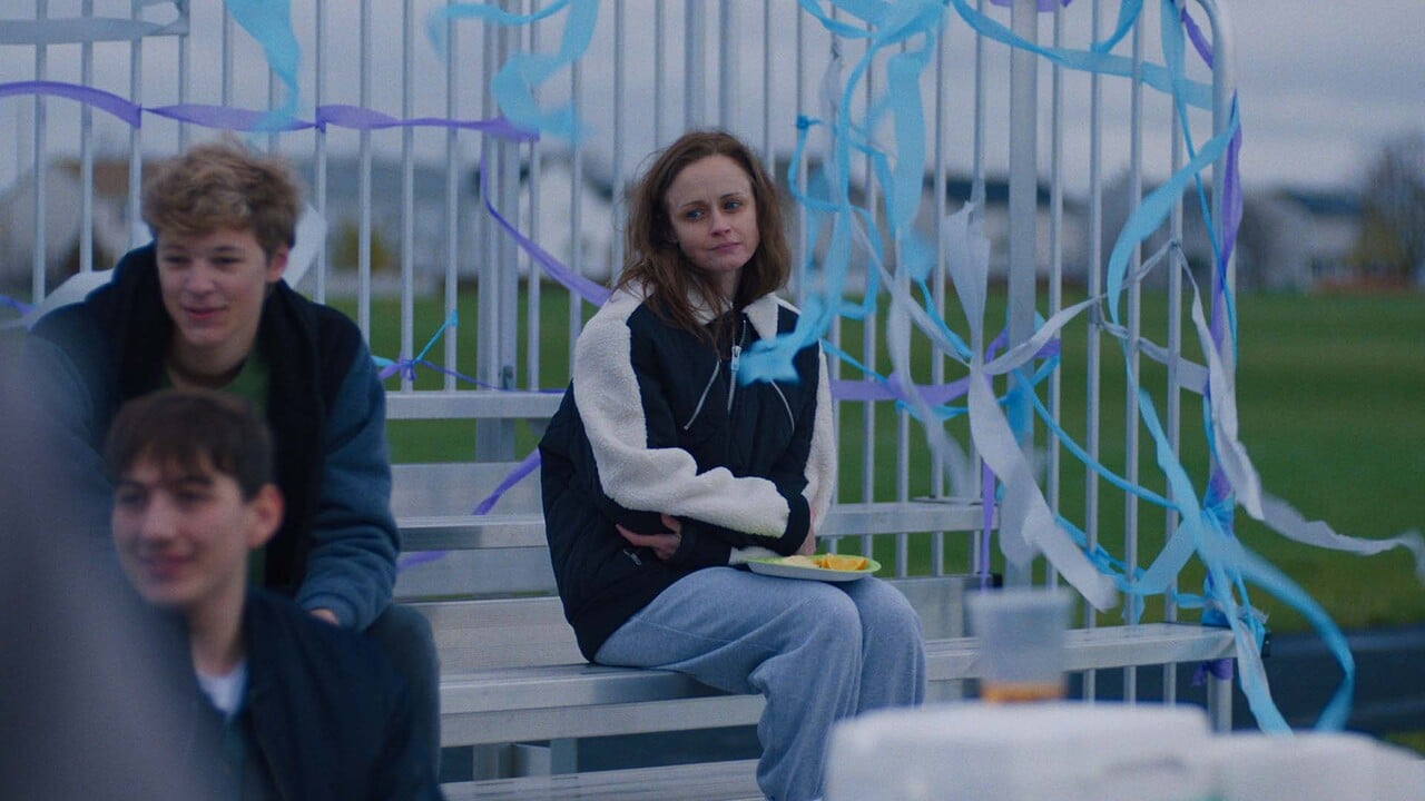 A young woman sits alone on bleachers decorated with blue streamers, holding a plate, while a few others sit nearby on an overcast day.