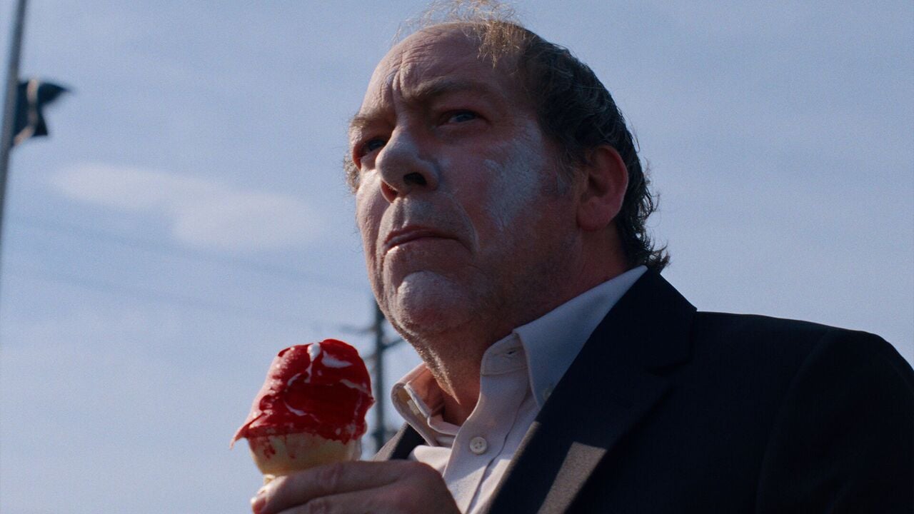 A man in a suit with white cream on his face holds a melting ice cream cone with red topping, standing outdoors under a blue sky.