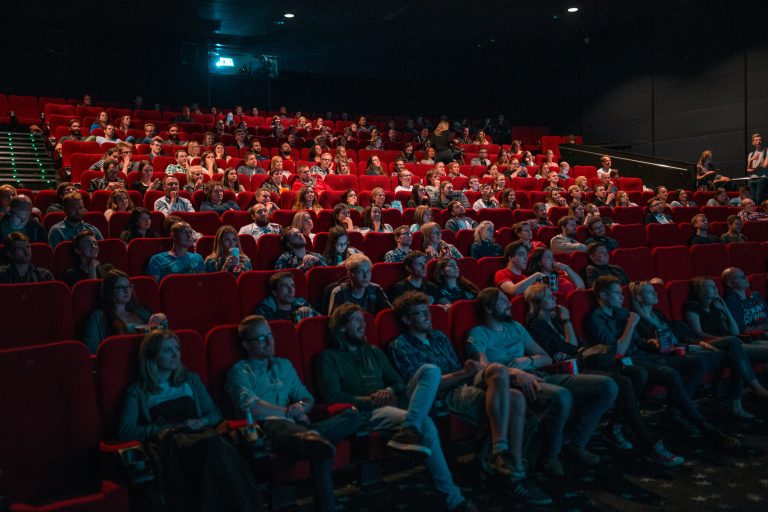 Why We Can’t Stop Ranking Films, Series, And Geek Culture A full audience sits in red theater seats, attentively watching a movie on a large screen in a dimly lit cinema.