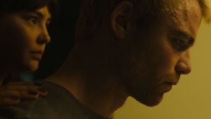 A woman stands behind a man, placing her hand on his shoulder as he looks forward with a serious expression in dim lighting.