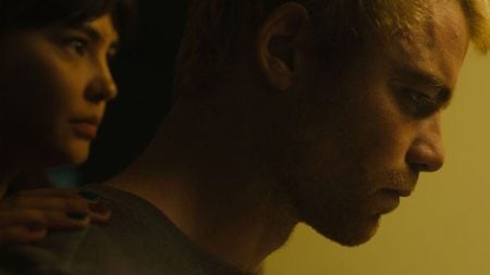 A woman stands behind a man, placing her hand on his shoulder as he looks forward with a serious expression in dim lighting.