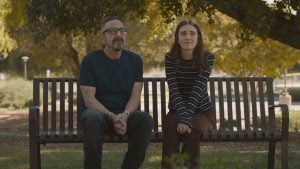 A man and a young woman sit side by side on a bench in a park, both looking ahead with neutral to thoughtful expressions. Trees and sunlight are visible in the background.