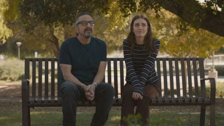 A man and a young woman sit side by side on a bench in a park, both looking ahead with neutral to thoughtful expressions. Trees and sunlight are visible in the background.
