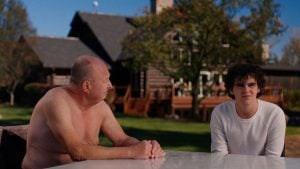 An older shirtless man and a younger man in a white shirt sit at an outdoor table in front of a large house on a sunny day.