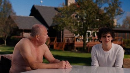 An older shirtless man and a younger man in a white shirt sit at an outdoor table in front of a large house on a sunny day.
