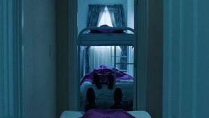 A dimly lit room with two beds and a bunk bed, each occupied by a person lying down covered in purple sheets. Light filters through curtained windows in the background.