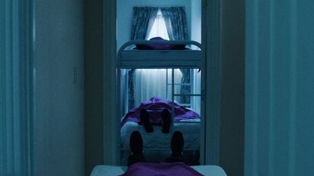 A dimly lit room with two beds and a bunk bed, each occupied by a person lying down covered in purple sheets. Light filters through curtained windows in the background.
