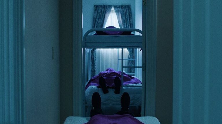A dimly lit room with two beds and a bunk bed, each occupied by a person lying down covered in purple sheets. Light filters through curtained windows in the background.