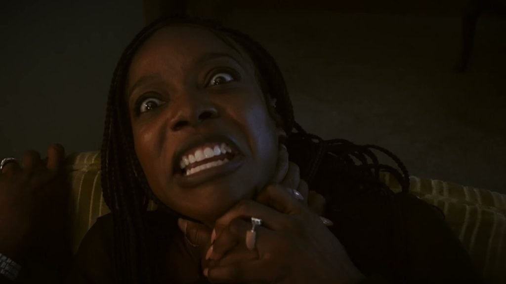 A woman with braided hair grimaces and holds her hands to her throat as if being choked, showing fear and distress.