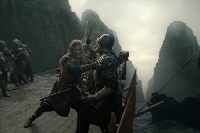 A bearded warrior in medieval armor fights a knight on a wooden bridge surrounded by rocky cliffs, while others battle in the background under a cloudy sky.