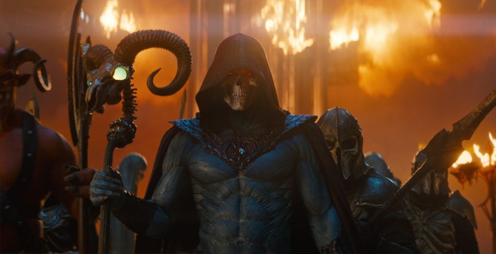 A hooded, skeletal figure with glowing red eyes and blue skin holds a staff, flanked by armored beings, with fire and smoke in the background.
