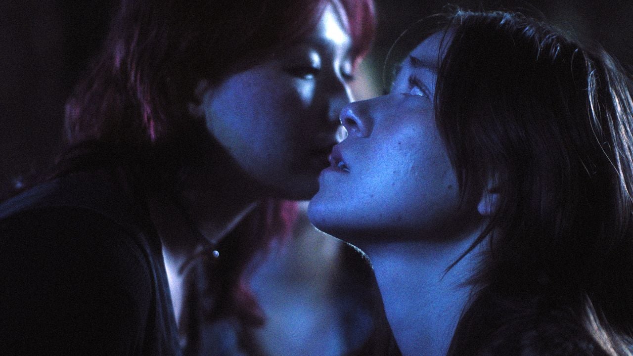 Two women in dim blue lighting; one whispers into the other's ear while the second woman looks forward with a serious expression.