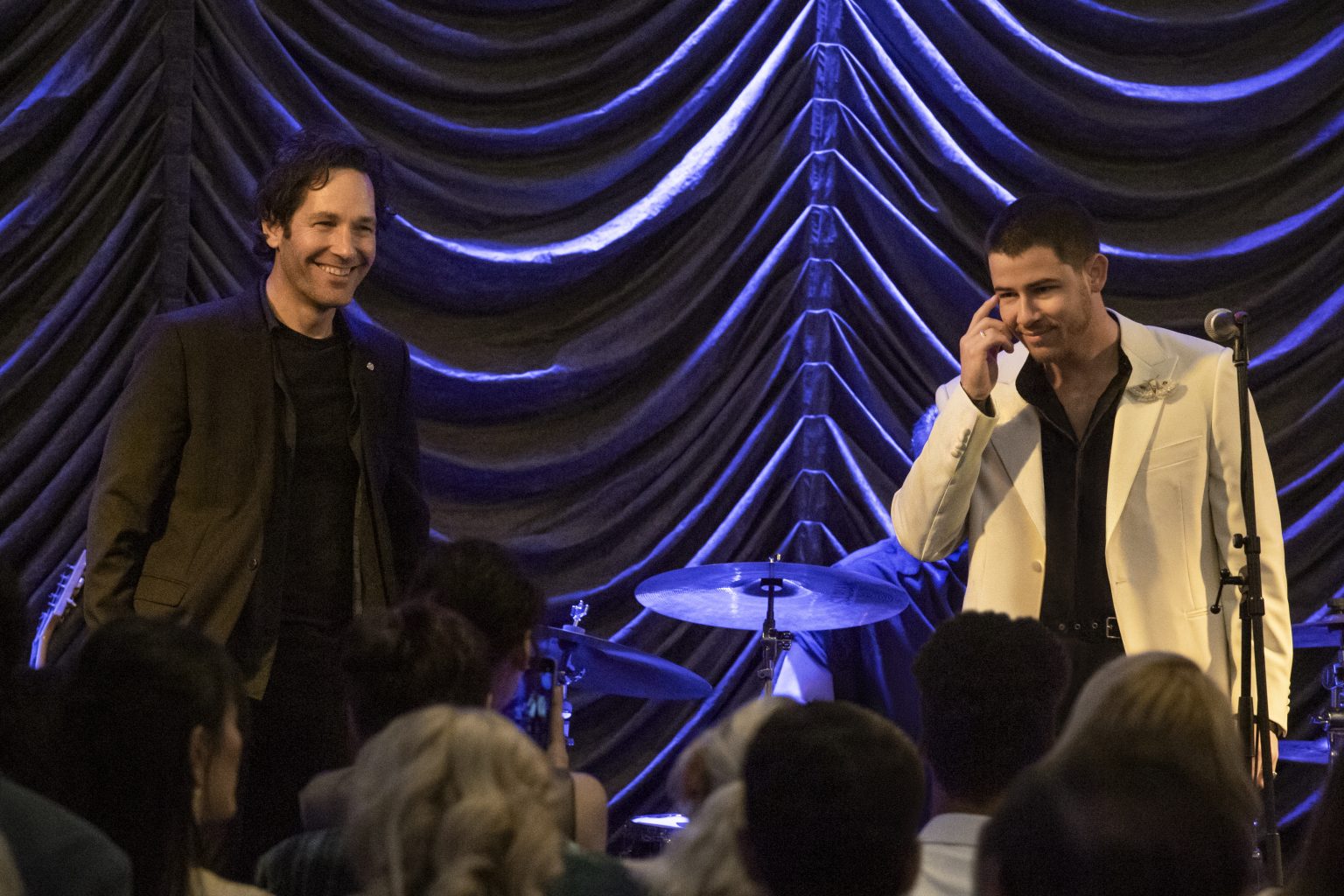 Two men stand on stage in front of a drum set and gathered audience, one wearing a dark jacket and the other a white suit jacket, with pleated curtains in the background.