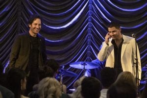 Two men stand on stage in front of a drum set and gathered audience, one wearing a dark jacket and the other a white suit jacket, with pleated curtains in the background.