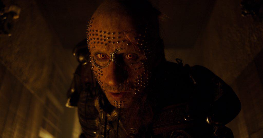 A person with a shaved head and face covered in metal studs stares intently, dressed in dark, armored clothing in a dimly lit setting.