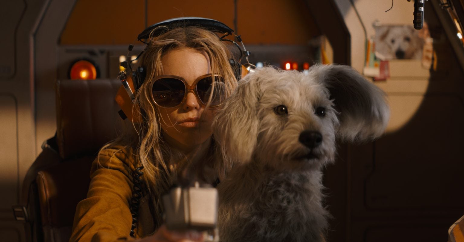 A woman wearing large sunglasses and headphones sits in a cockpit, holding a control lever, with a fluffy gray dog in the foreground.